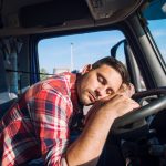 A tired truck driver wearing a red plaid shirt rests his head on the steering wheel inside the truck cabin during the daytime. The image highlights the importance of driver rest and fatigue management for road safety. The background shows a parking lot with other trucks, suggesting a rest stop or break area.