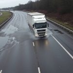 A white cargo truck is seen traveling on a wet highway, creating water splashes as it moves. The road appears damp, possibly after recent rain, with puddles scattered across the lanes. The surrounding environment consists of grassy areas and trees lining the highway. The sky is overcast, suggesting cloudy or rainy weather. The highway curves in the distance with a few other vehicles visible further down the road.