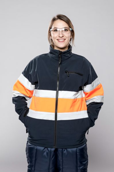 Smiling woman wearing safety glasses and a high-visibility jacket with reflective stripes, ready for work in a safety-conscious environment.